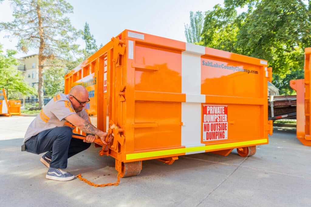 A man is tying a rope to an orange dumpster.